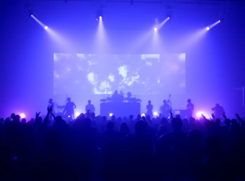 A stage bathed in blue light, with silhouetted figures performing electronic music in front of a large screen displaying abstract visuals.